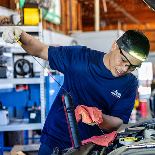 A mechanic in a blue shirt, safety glasses, and headlamp checks a car's oil level with a dipstick.