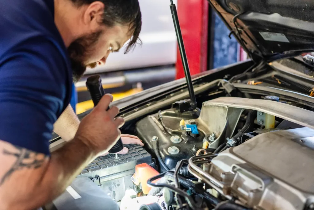 A bearded mechanic in a blue shirt inspects a car engine with a flashlight, focusing on details.